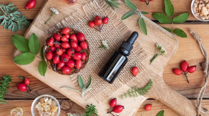 Flores, frutos e sementes de rosa mosqueta dispostos numa mesa ao lado de um frasco de sérum