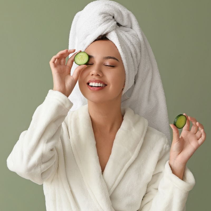 modelo com roupão e toalha enrolada em cabelo segurando um pepino em frente ao rosto