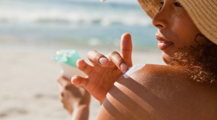 Foto de uma modelo na praia aplicando protetor solar em creme no ombro.