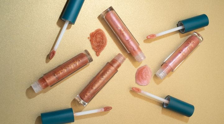 Quatro embalagens de gloss labial aberta, em tom rosê sobre um apoio areia
