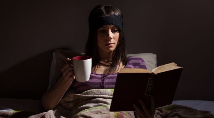 mulher deitada na cama segurando uma xicará de chá e lendo um livro