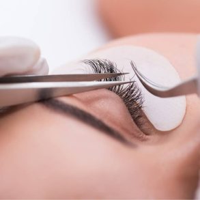 Close-up de um olho durante a aplicação de extensões de cílios, com pinças e adesivos protetores visíveis.