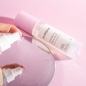 pessoa espirrando a bruma facial em um espelho deitado em um fundo rosa vazio